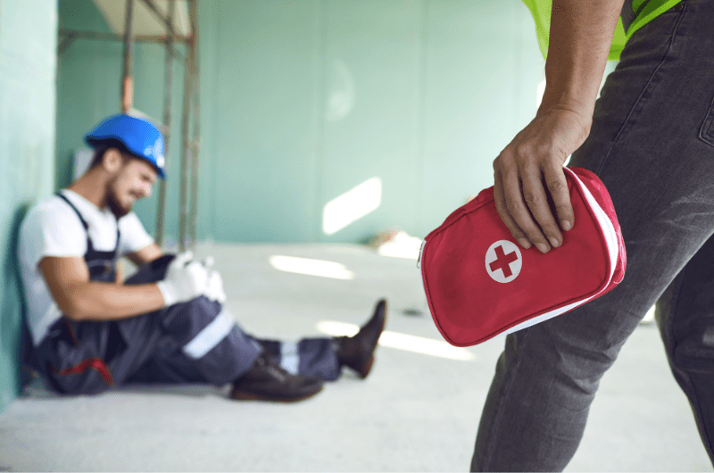 A construction worker is on the ground in pain after falling, and his co-worker is coming to help with a first aid kit